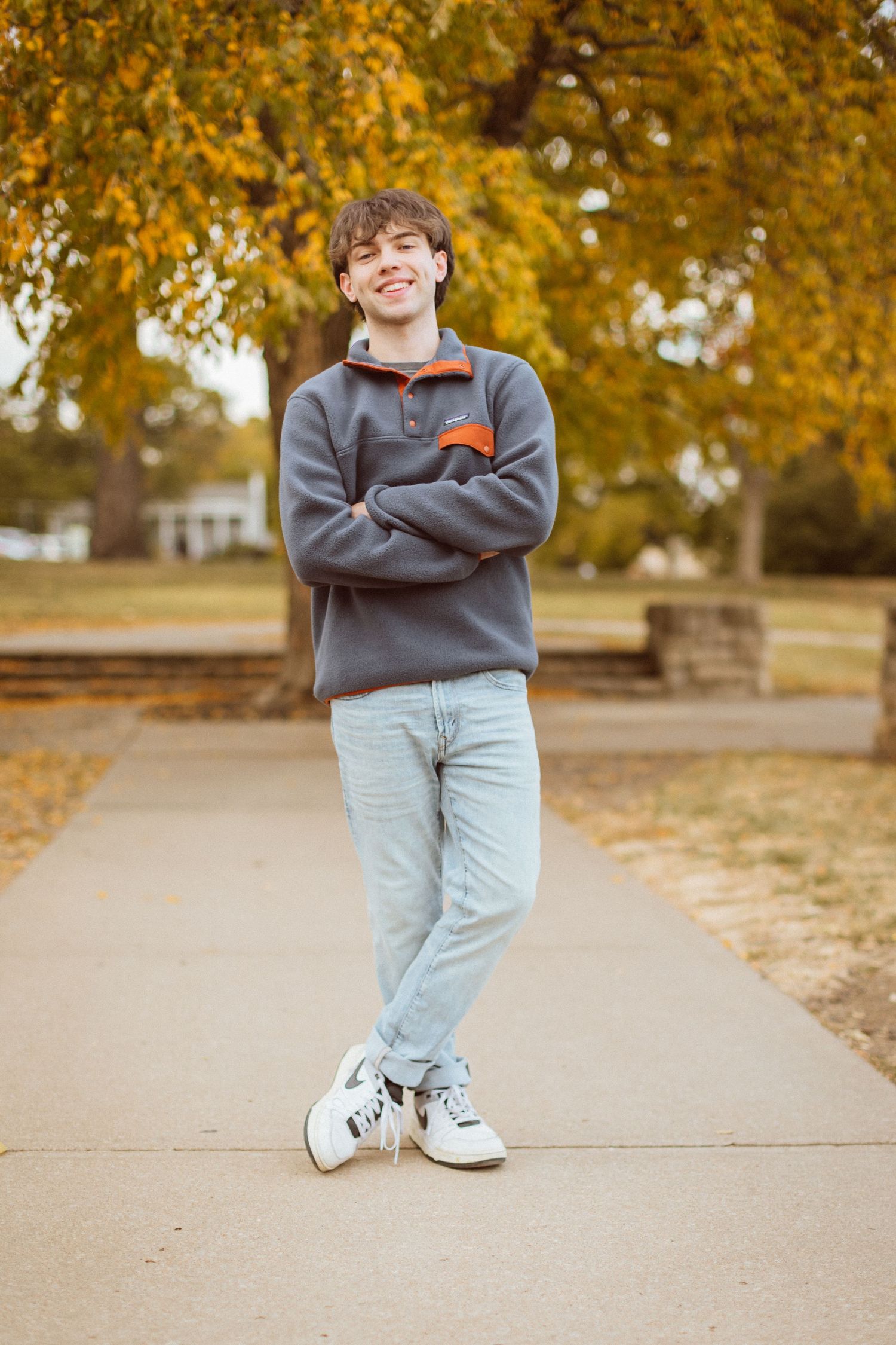 A young man stands with arms and ankles crossed in front of colorful fall trees during his Kansas City senior photo session.