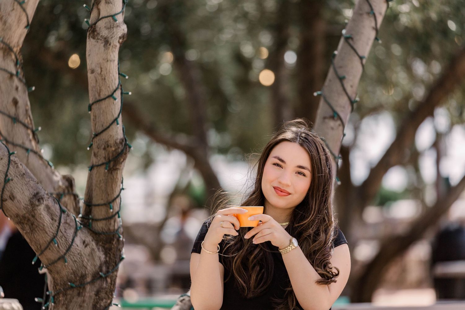 A woman in a black dress poses thoughtfully against a tree trunk in an outdoor garden setting.