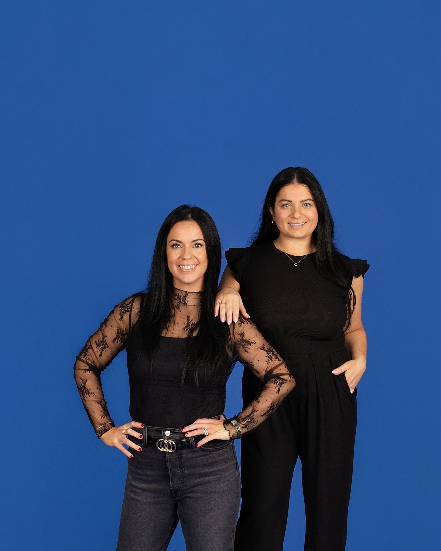 Two people in black outfits pose together against a bright blue backdrop in a studio setting.