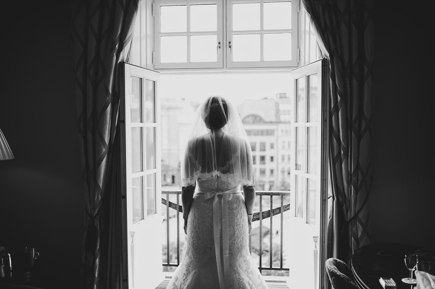 A silhouetted figure stands at an open window wearing a flowing dress in black and white photography.