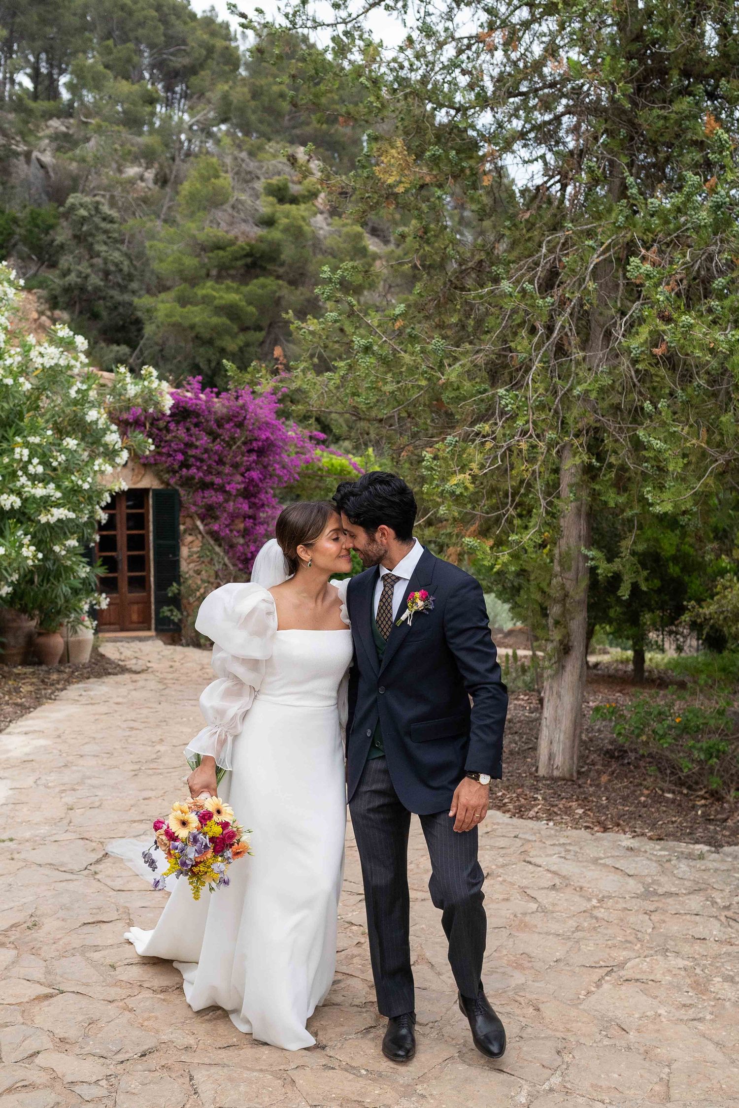 A romantic wedding couple walking along a stone path surrounded by blooming purple bougainvillea and lush greenery.