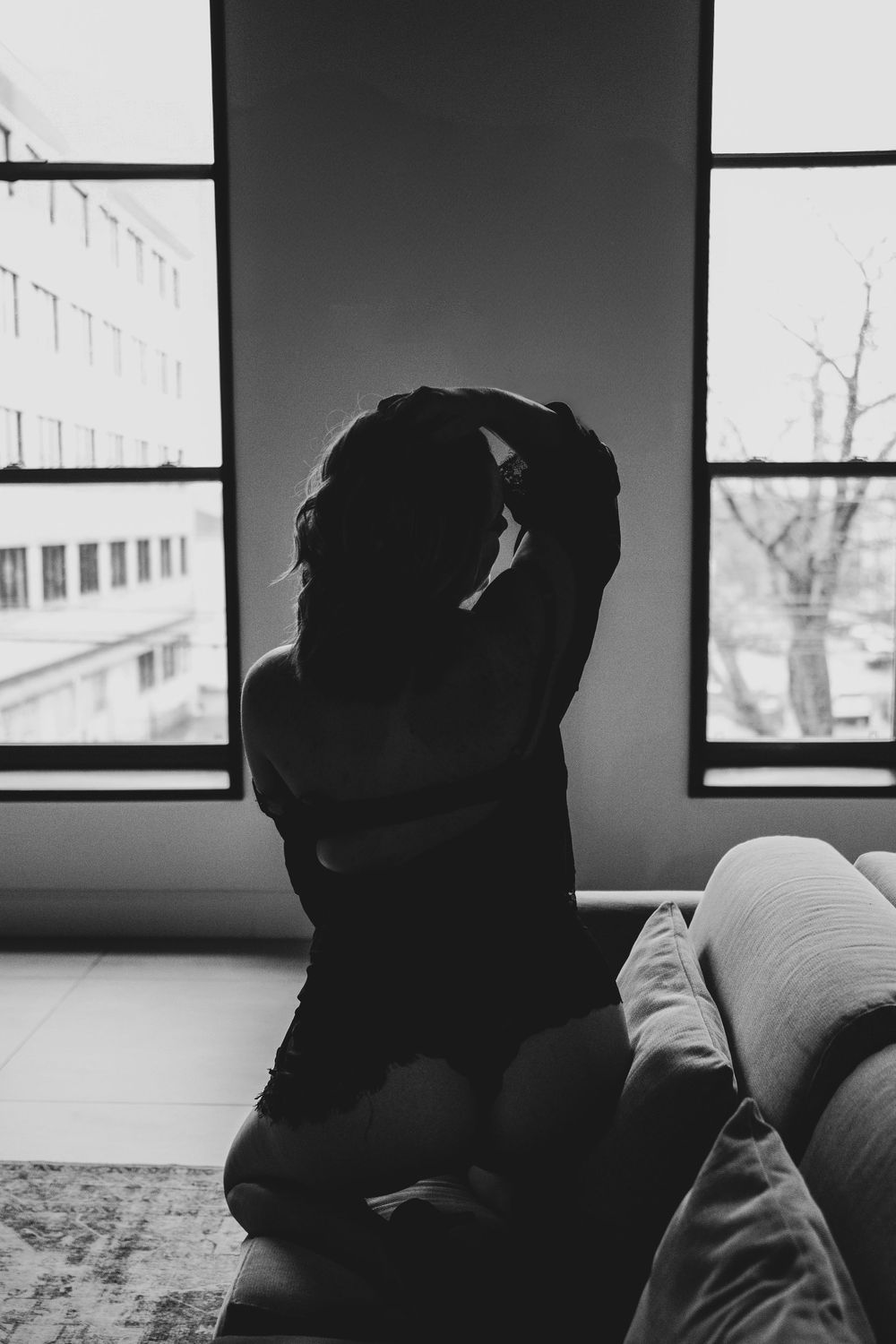 Black and white silhouette of woman sitting by large windows adjusting her hair.