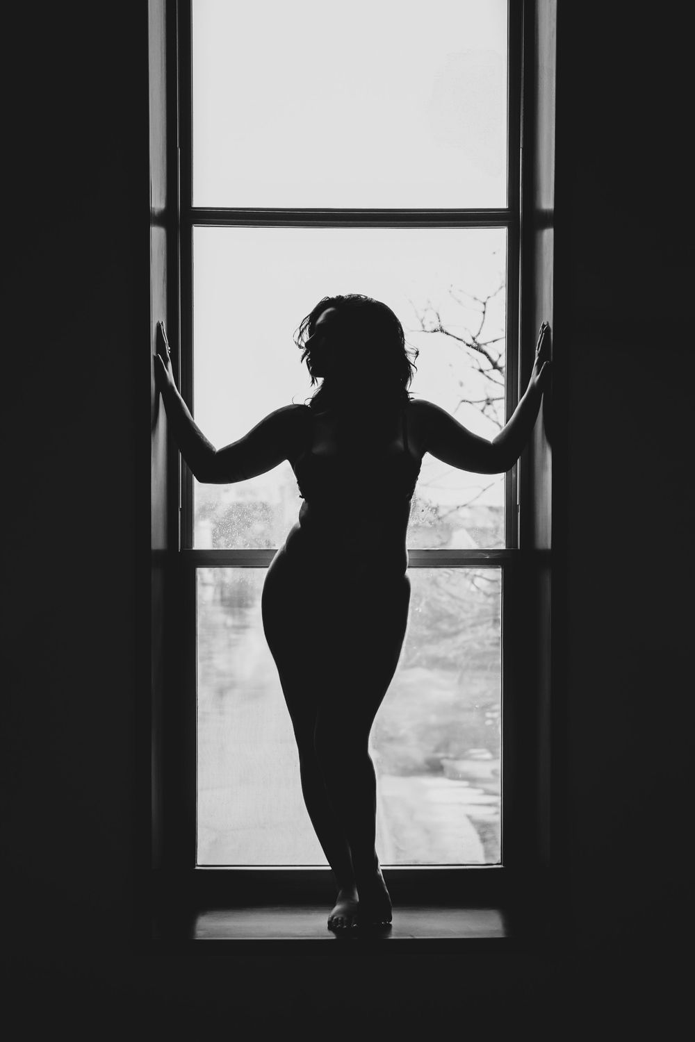 Black and white silhouette of woman standing at large window with hands on frame.