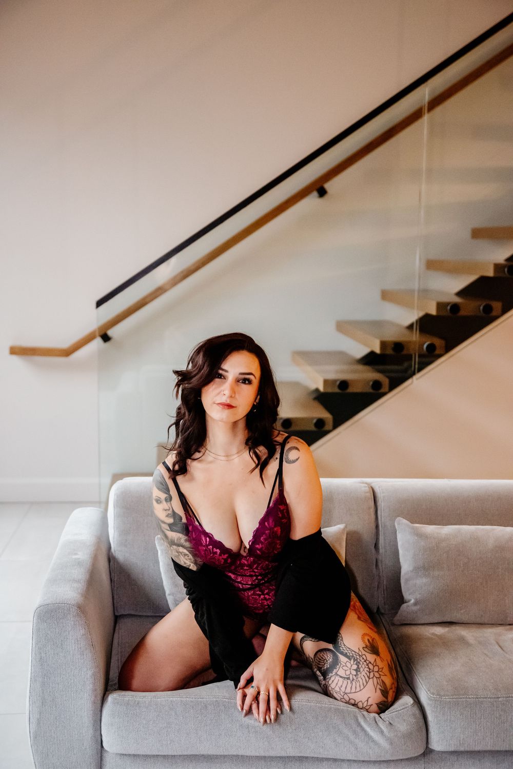 Tattooed brunette woman in burgundy lace lingerie sitting on gray sofa near modern staircase.