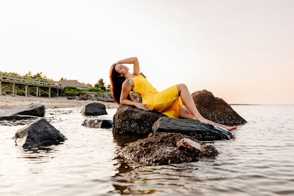 Woman in yellow dress posing on rocks by the water at sunset.