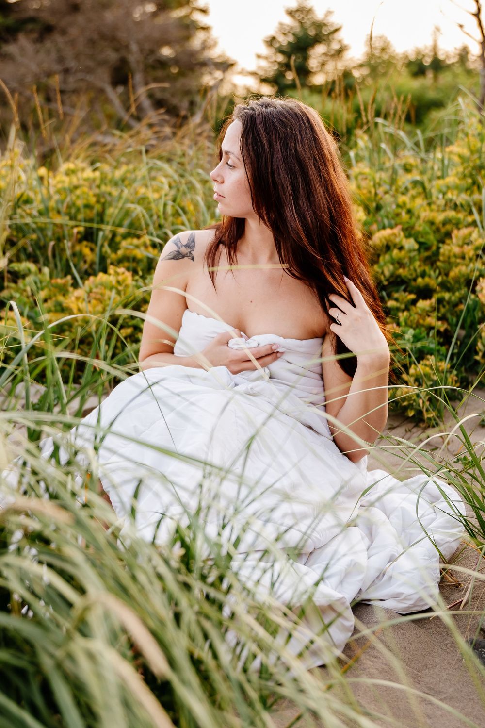 Woman in white wrap sitting among tall green grass at golden hour.