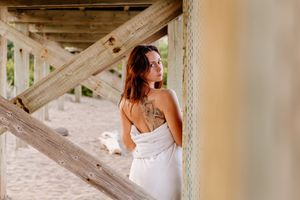 Woman in white strapless dress with tattoo standing under wooden beach structure.