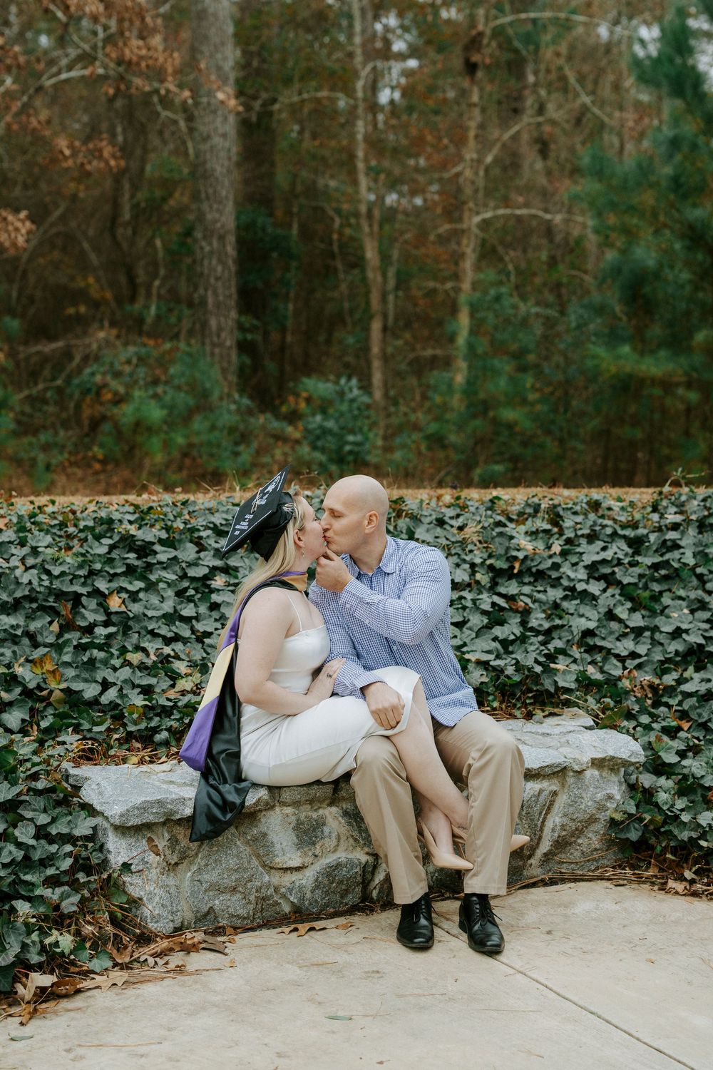 A couple shares a tender moment while sitting on a stone wall surrounded by ivy and autumn trees.