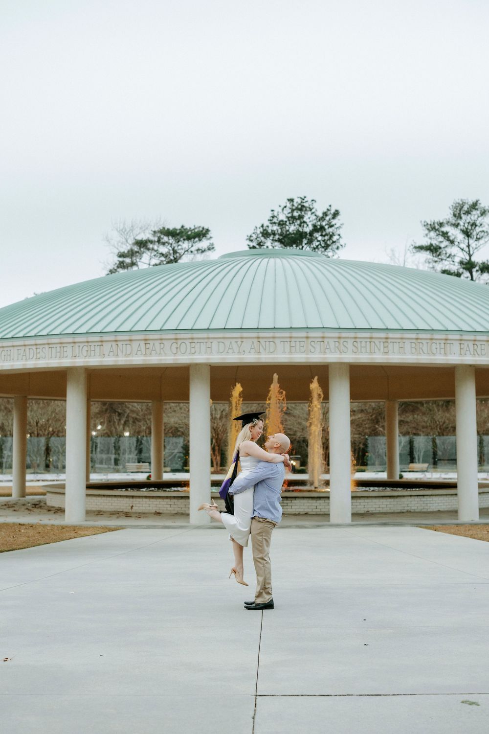 A couple performs a romantic dance lift pose under a gazebo with a green domed roof in a park setting.