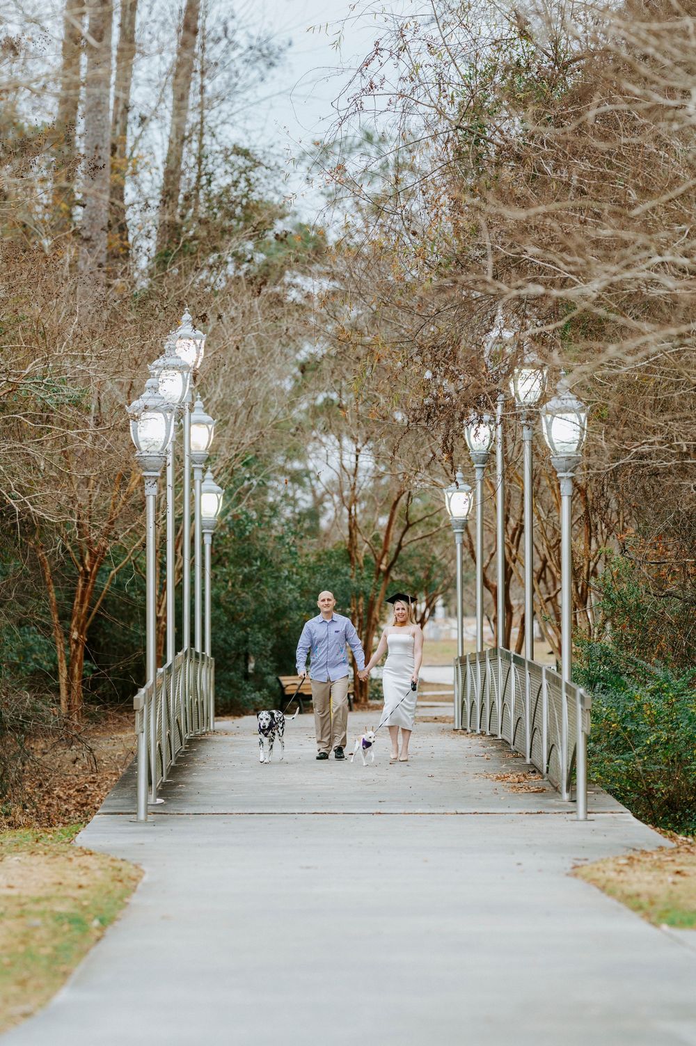 A couple walks along a scenic tree-lined pathway with white lamp posts in a park setting.