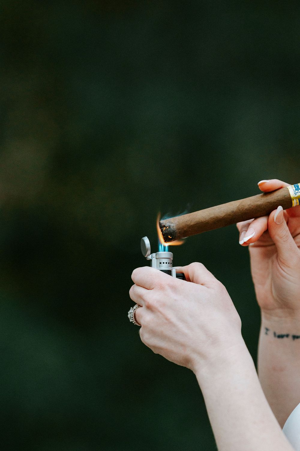 A person's hands light a cigar with a silver lighter against a dark background.
