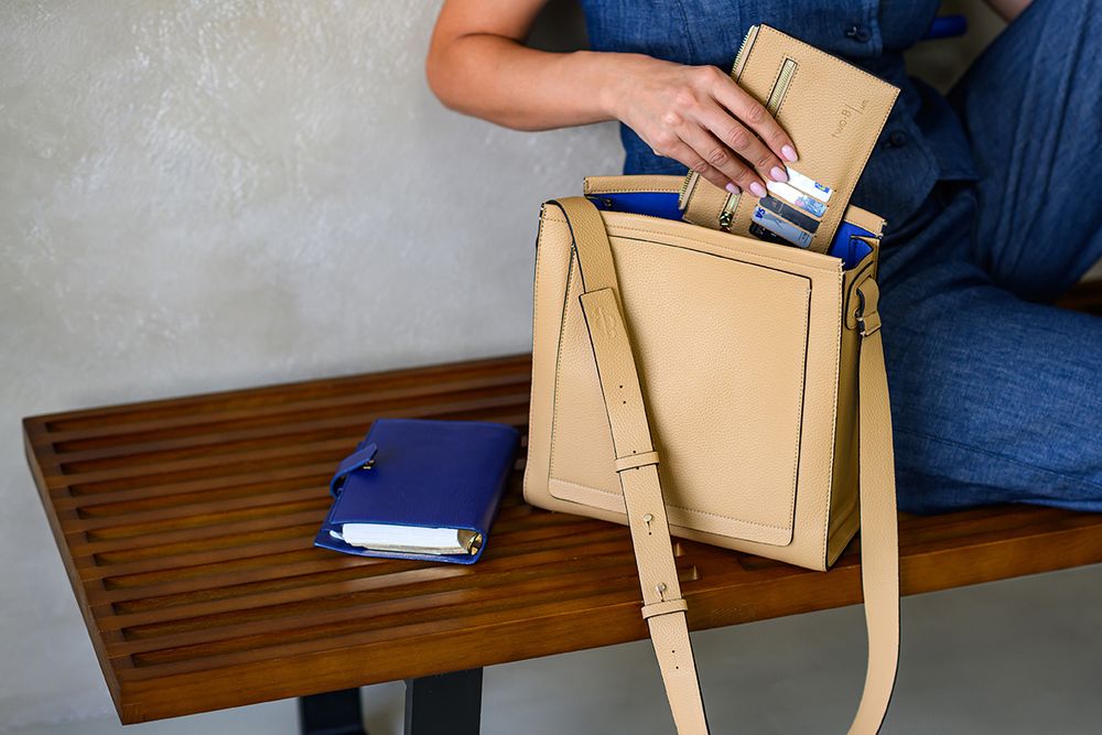 Hands holding and examining a beige leather handbag with structured design against blue background.