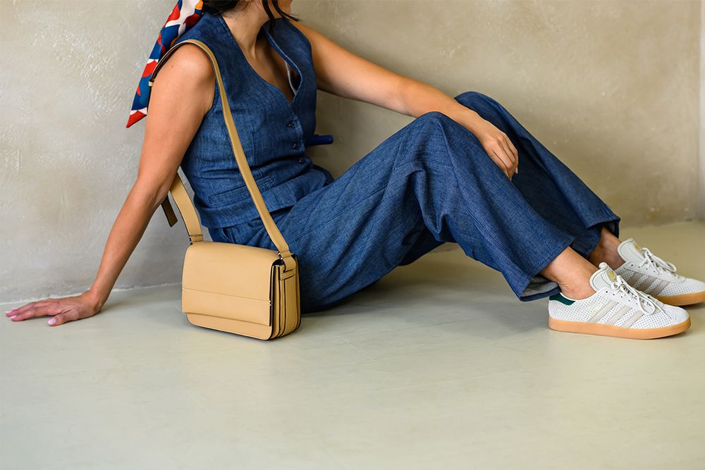 Person in casual denim outfit and white sneakers sitting on floor with tan crossbody bag.