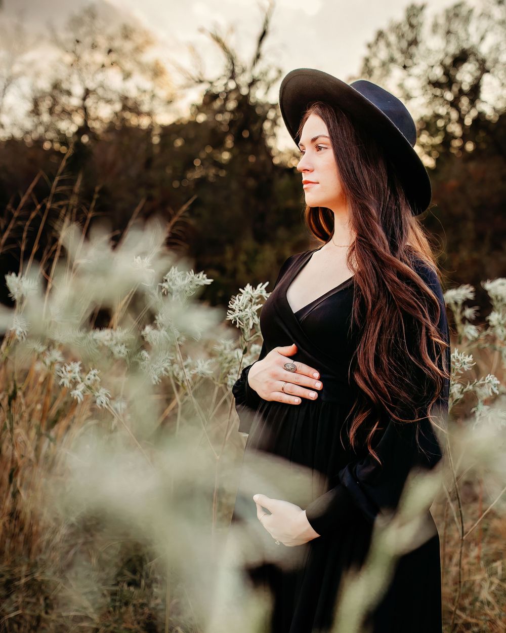 A woman in a black gown and wide-brimmed hat poses thoughtfully in a golden field at sunset in Fort Worth