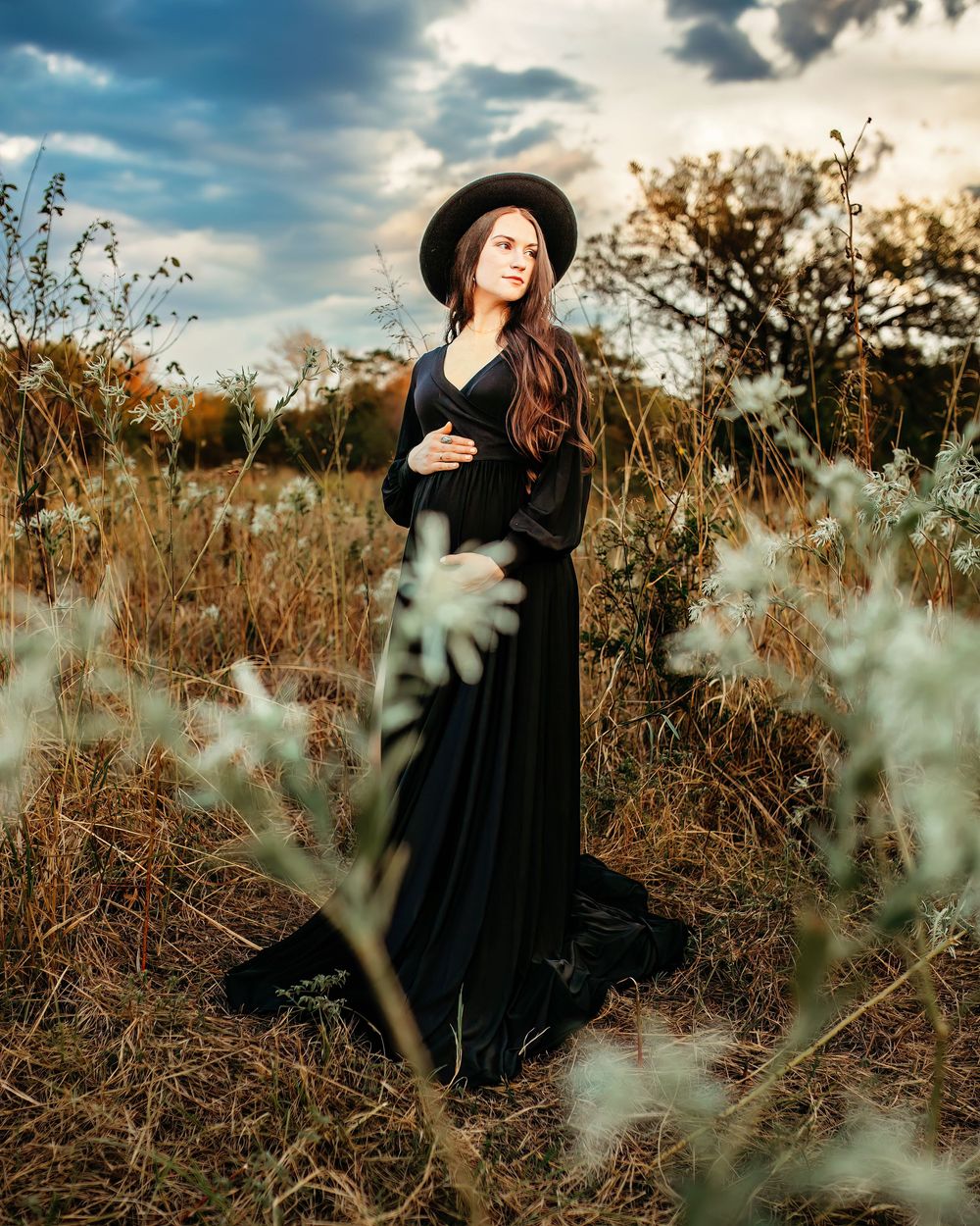A woman in a black dress and hat stands gracefully among tall autumn grasses against a moody cloudy sky in Fort Worth
