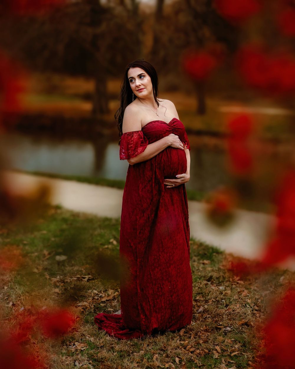A woman in a burgundy lace gown stands near a pond surrounded by autumn foliage with red bokeh effects in Colleyville, TX