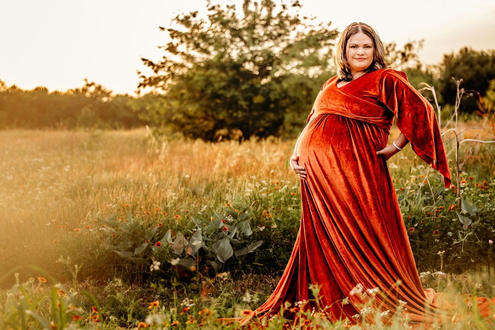 Pregnant woman in flowing rust colored velvet maternity gown posing in golden sunlit wildflower field in Fort Worth.