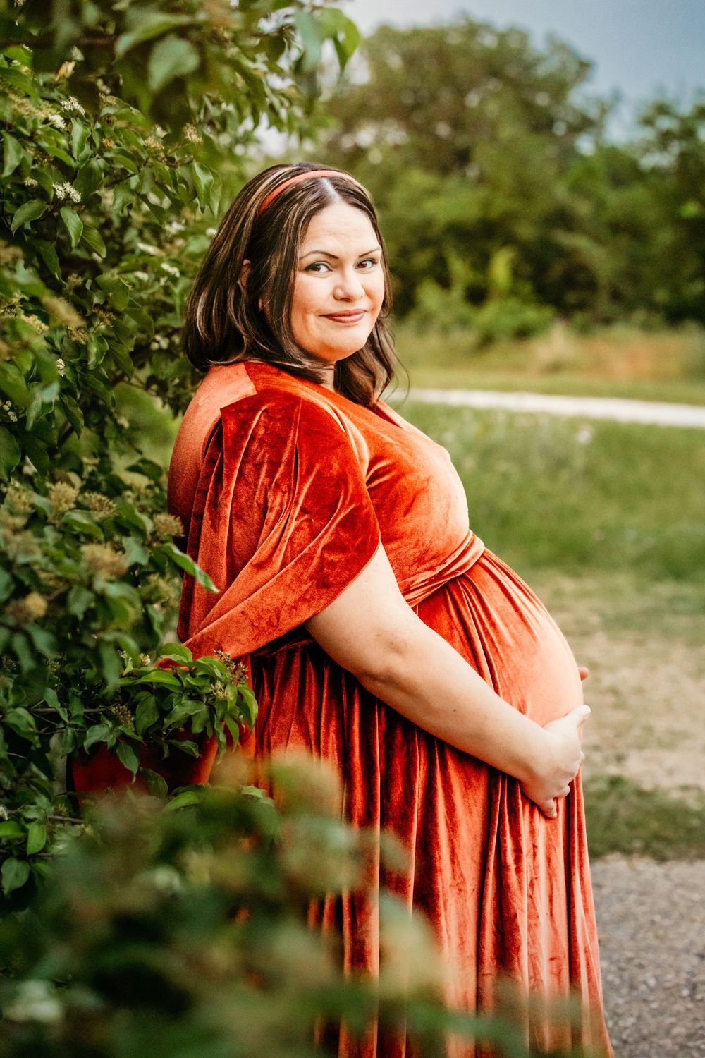 Pregnant woman in orange velvet dress posing outdoors among green bushes, cradling her baby bump with a gentle smile in Fort Worth, TX.
