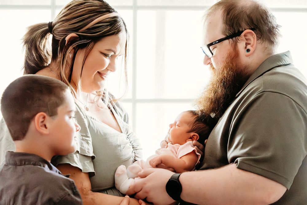 A family of four admires their newborn baby, with a bearded father and mother smiling warmly alongside their young son.