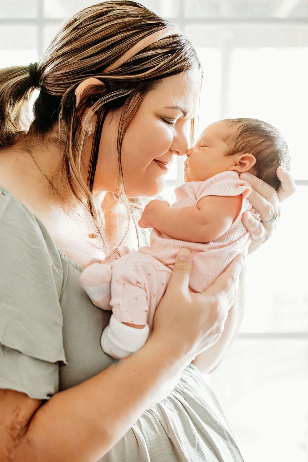 A mother tenderly touches noses with her newborn baby dressed in pink, bathed in soft natural light.