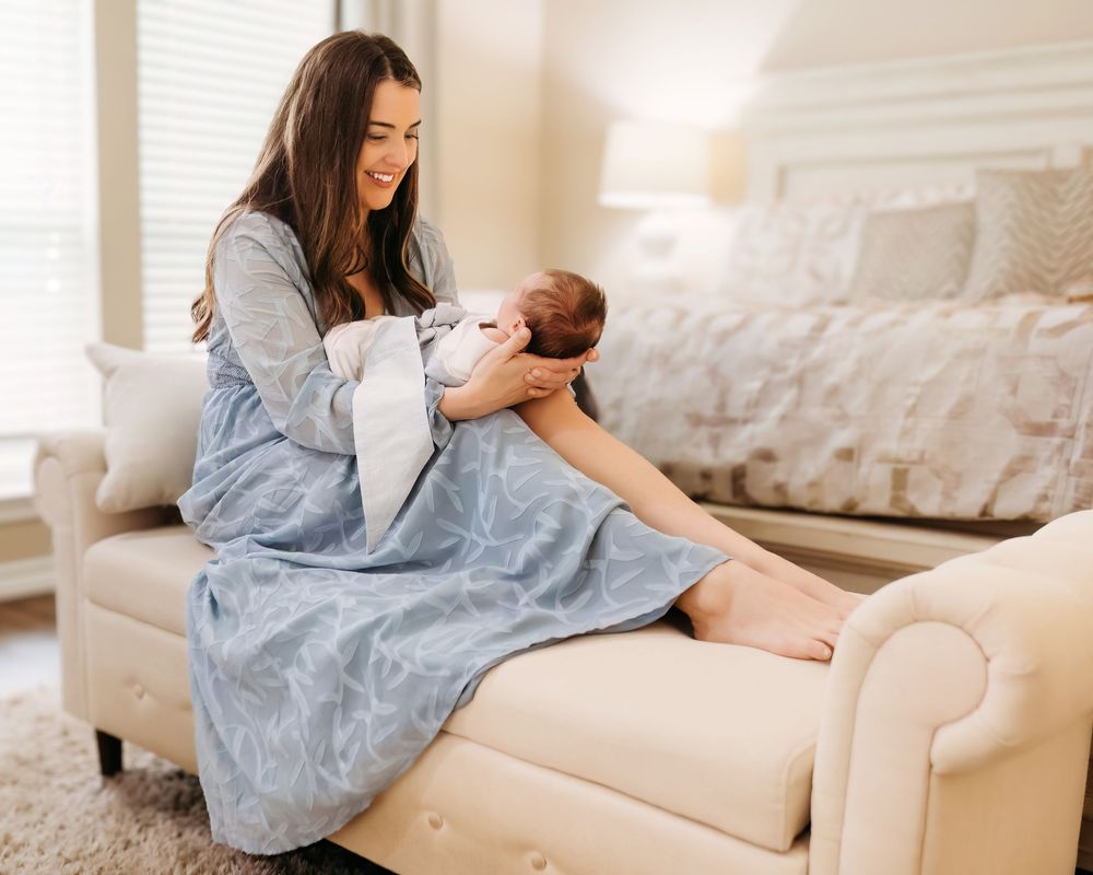 A mother in a blue dress sits on a white couch while bonding with her newborn baby in a bright room during in-home session in Southlake, TX
