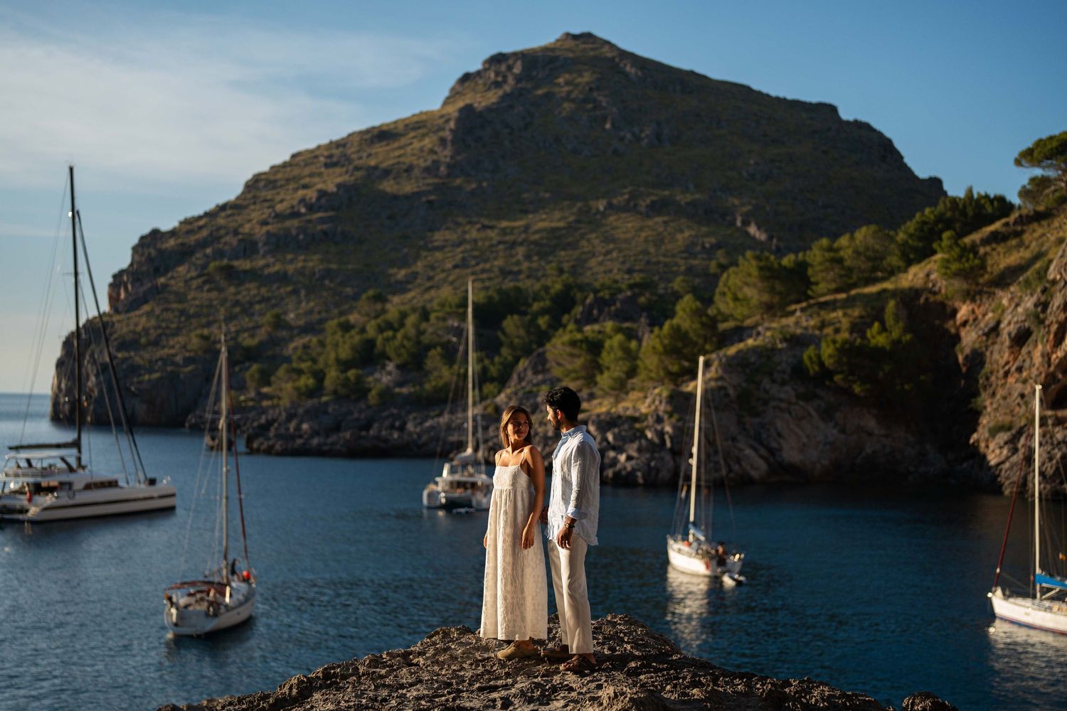 Sailboats anchored in a tranquil Mediterranean cove with rugged mountains and blue waters on a sunny day.