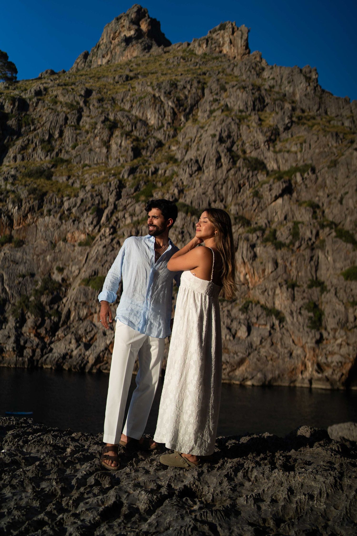 Couple in white outfits standing on rocky cliffs against deep blue sky in dramatic landscape.