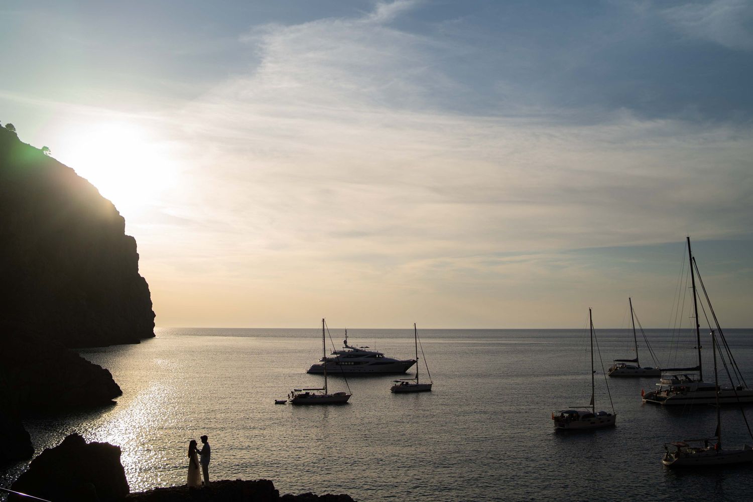 Sailboats anchored in a tranquil bay at sunset with silhouetted cliffs and golden sunlight reflecting on calm waters.