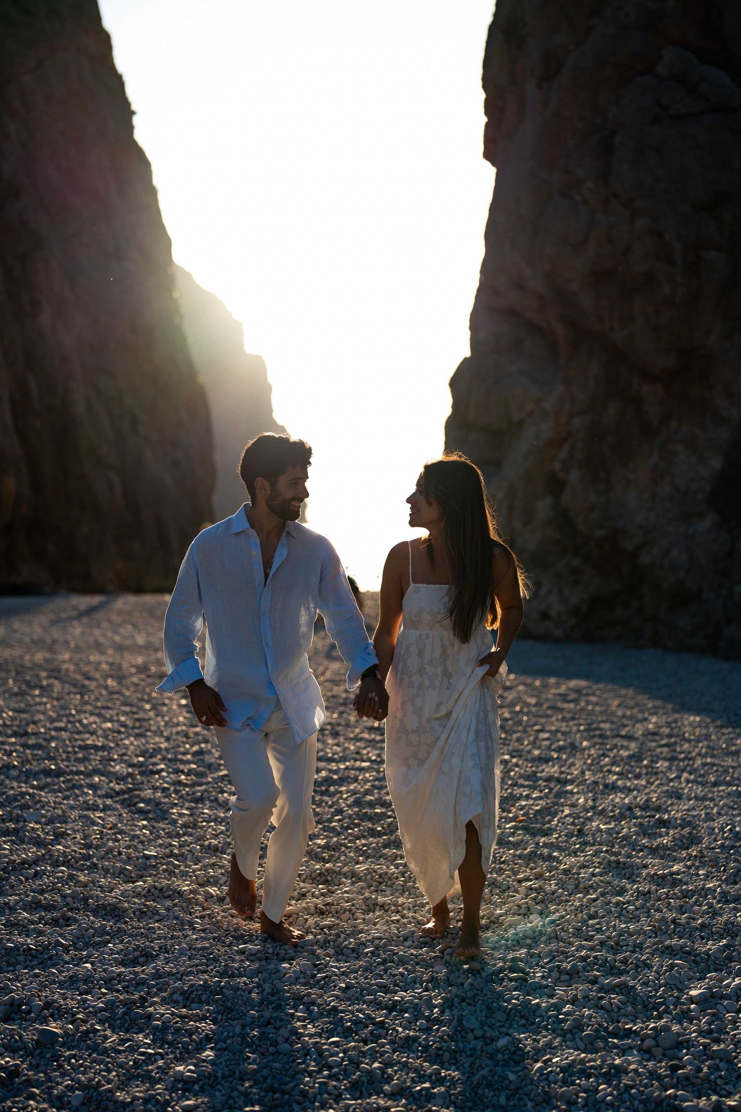Two people in white clothing walk along a rocky beach at sunset with towering cliffs behind them.