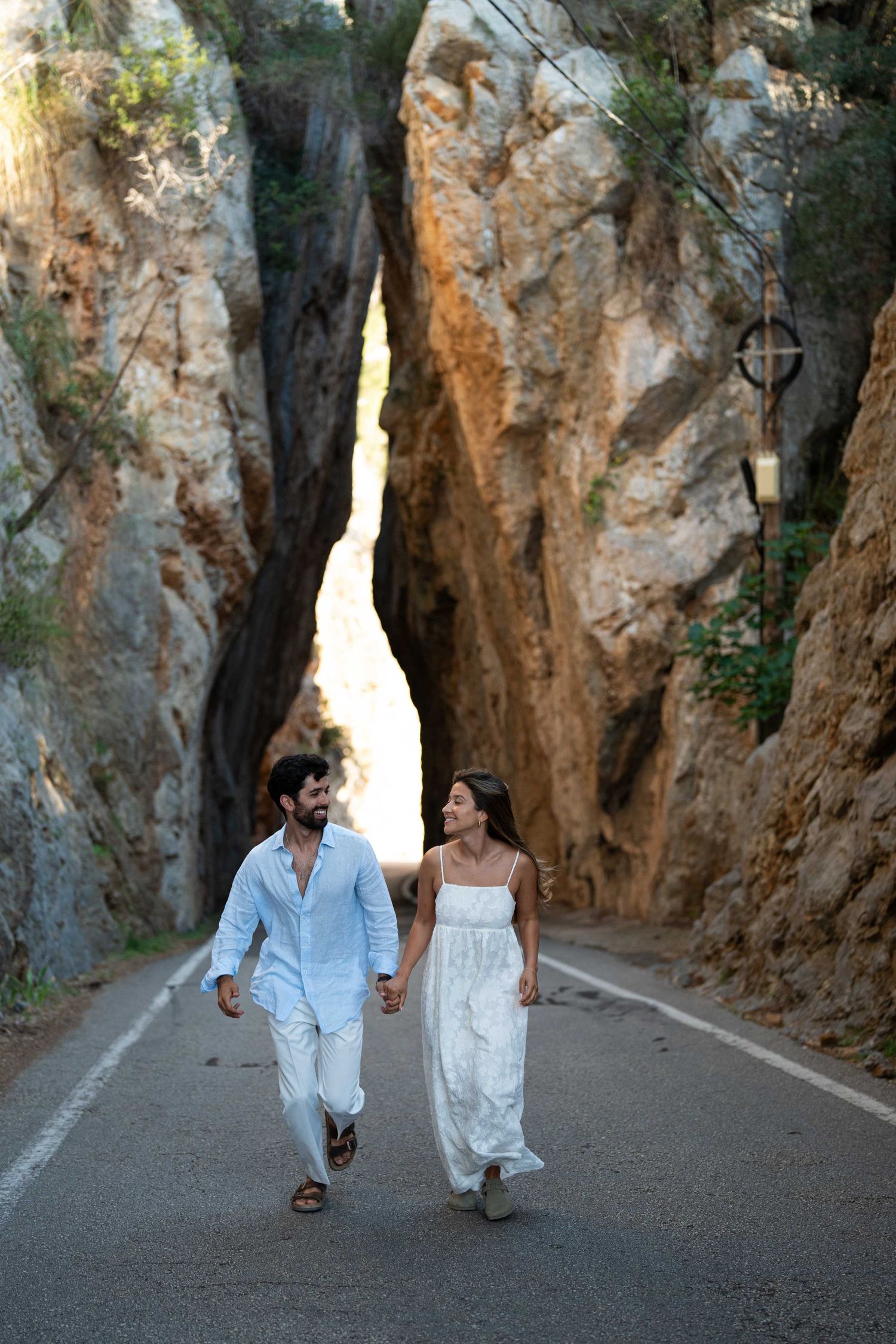A couple walks hand in hand down a scenic road between towering limestone cliffs in summer attire.