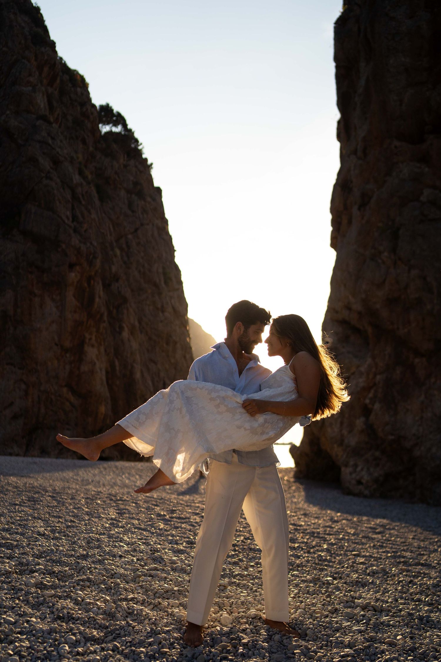 Romantic silhouette of couple embracing at sunset between dramatic rocky cliffs on a beach.