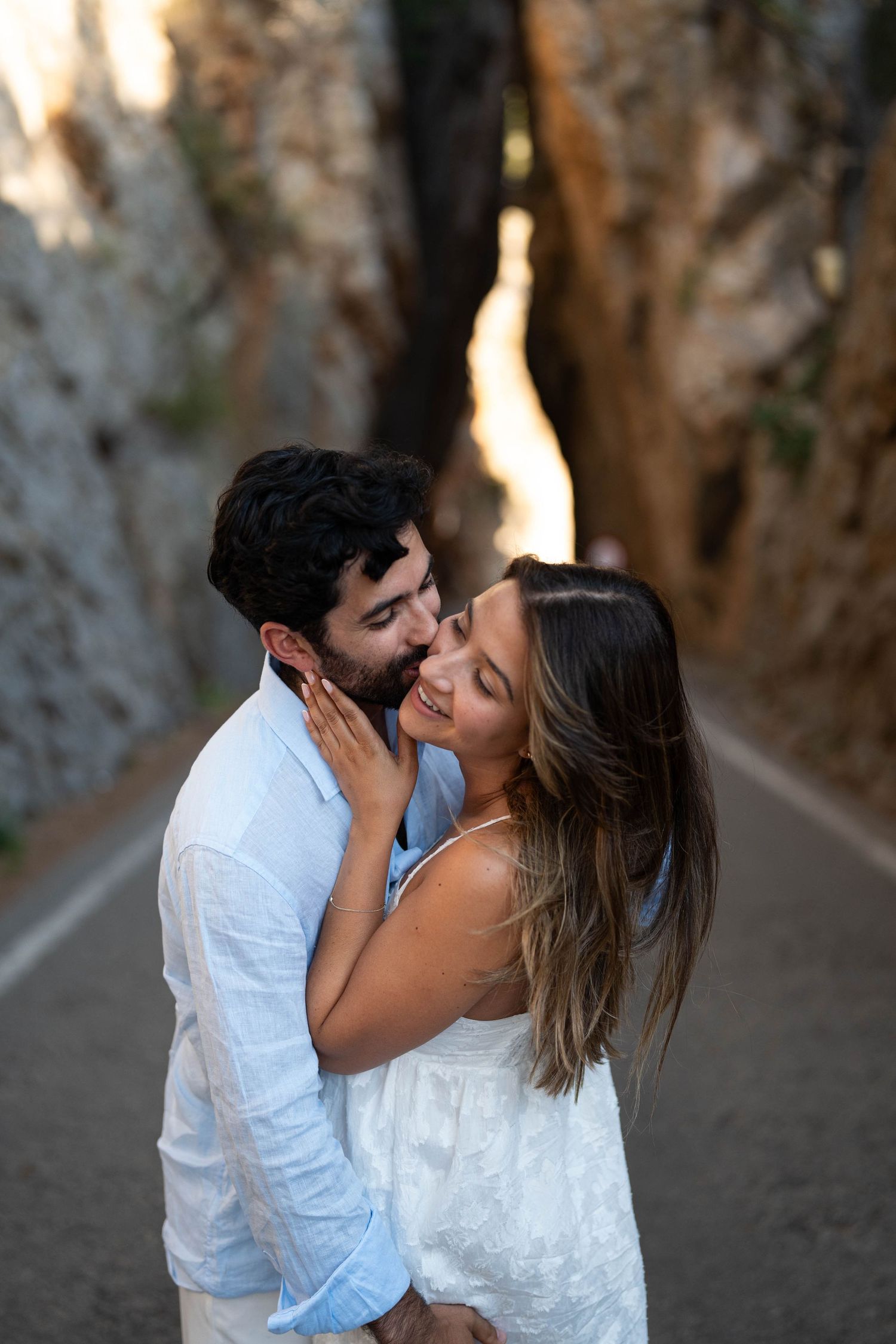 A romantic couple in white outfits share an intimate moment on a tree-lined path at sunset.