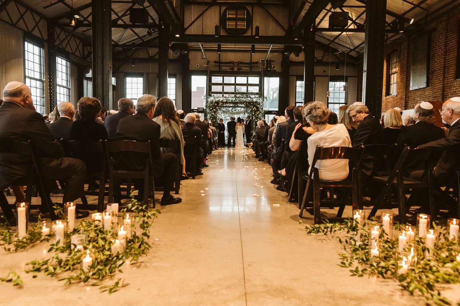 Panoramic view of a rustic indoor wedding ceremony with candlelit aisle decorated with greenery and flowers.