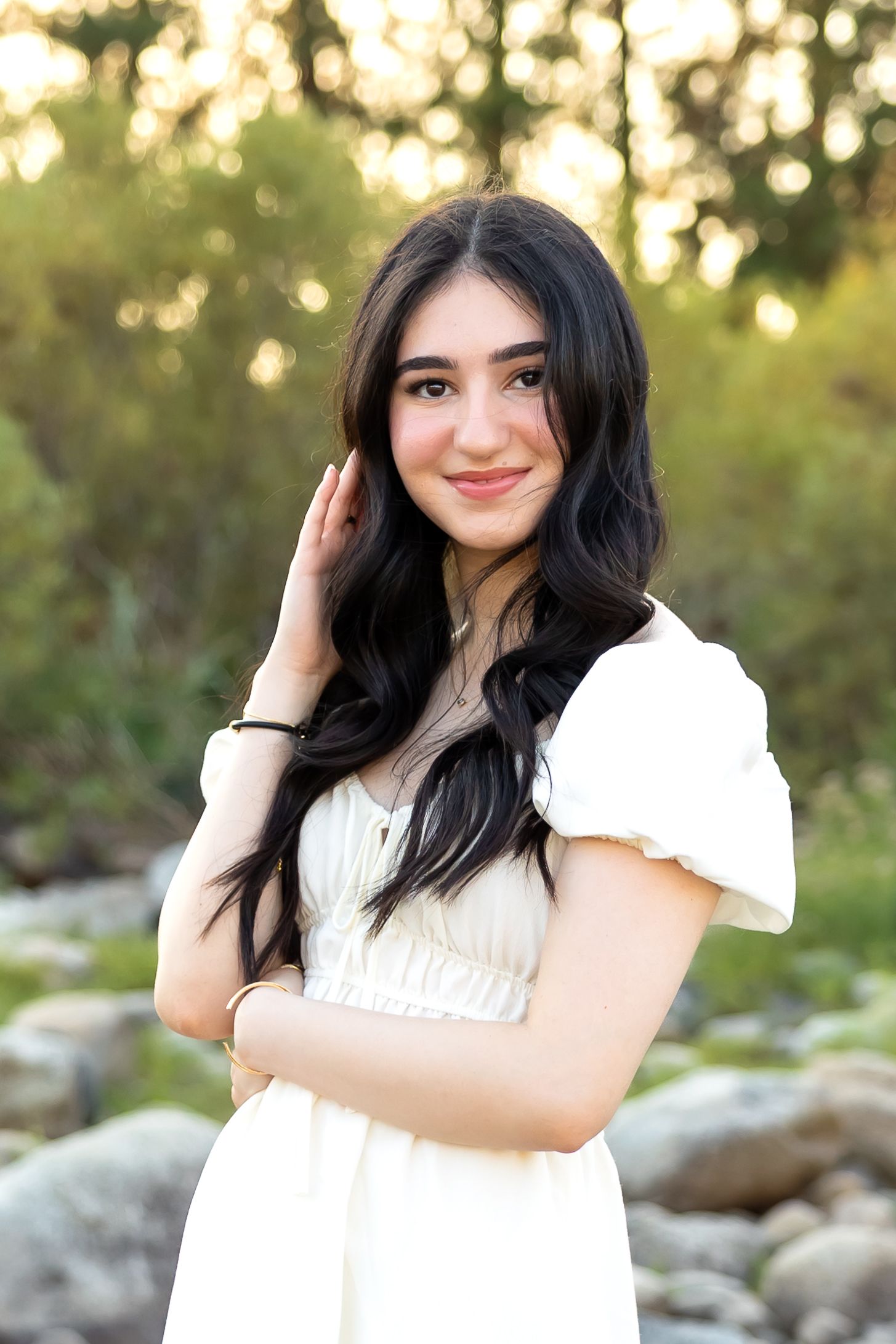A person in a white dress poses outdoors against a natural backdrop with bokeh lighting effects at sunset.