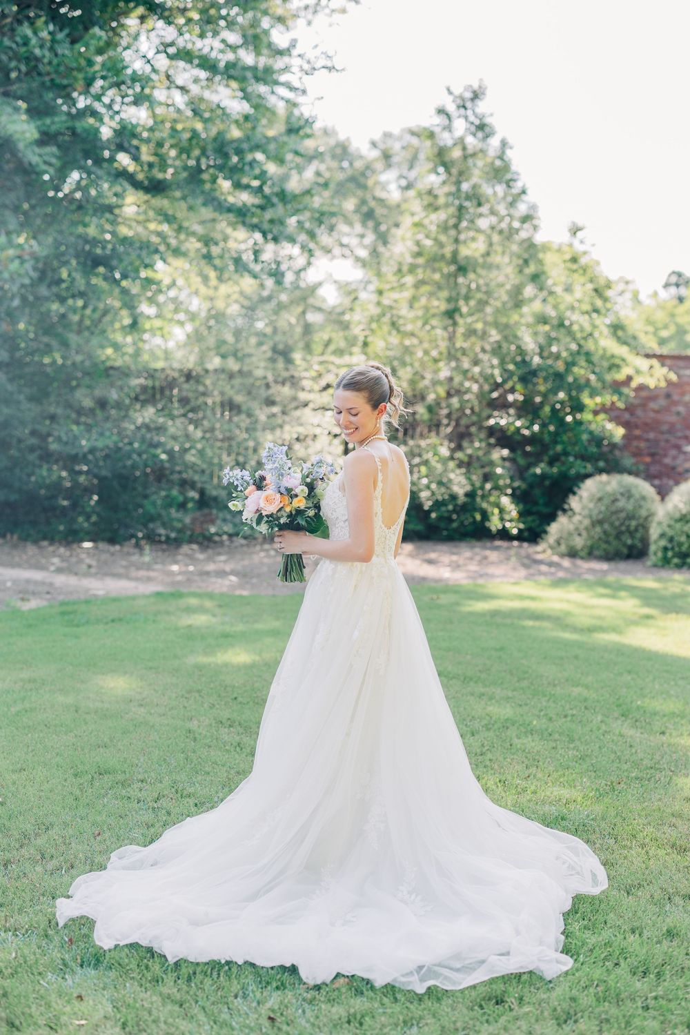 A bride in a flowing white gown with a long train holds a colorful bouquet while standing on a sunlit garden lawn at The Carl House