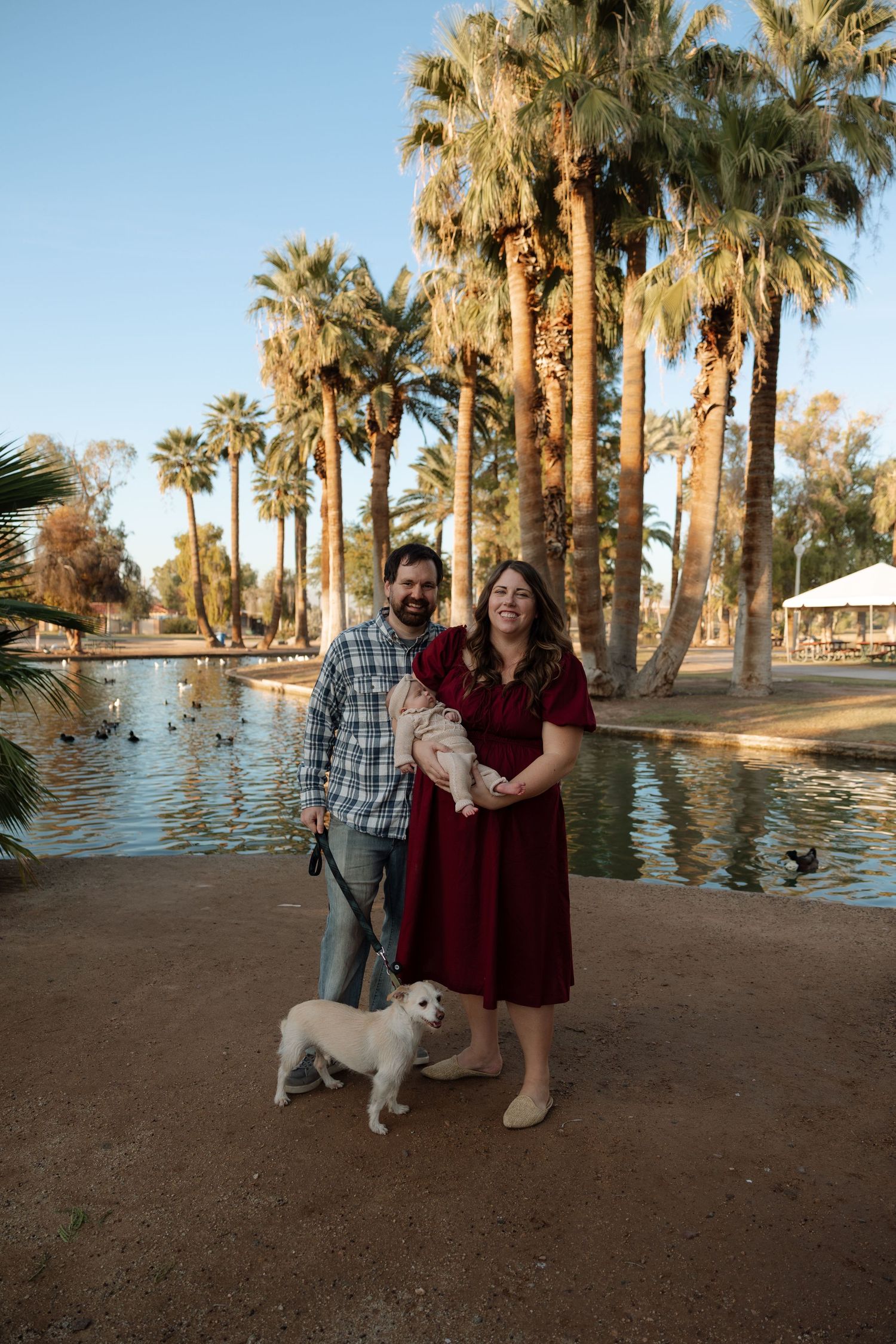 A couple poses with their white dog near palm trees and water during golden hour at a desert oasis.