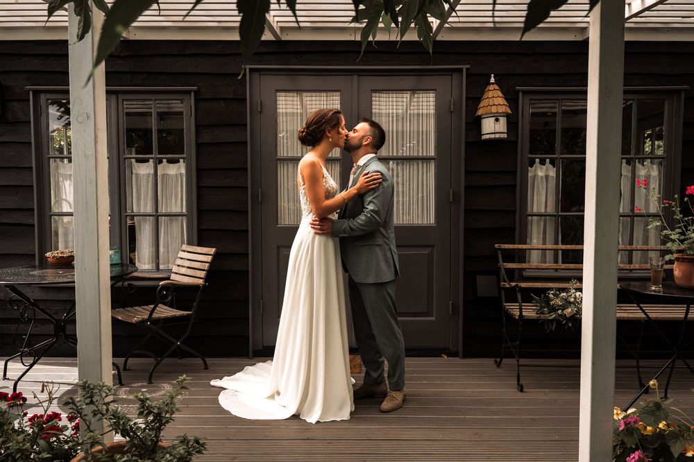 Newlyweds share a romantic moment on a front porch with white columns and greenery.