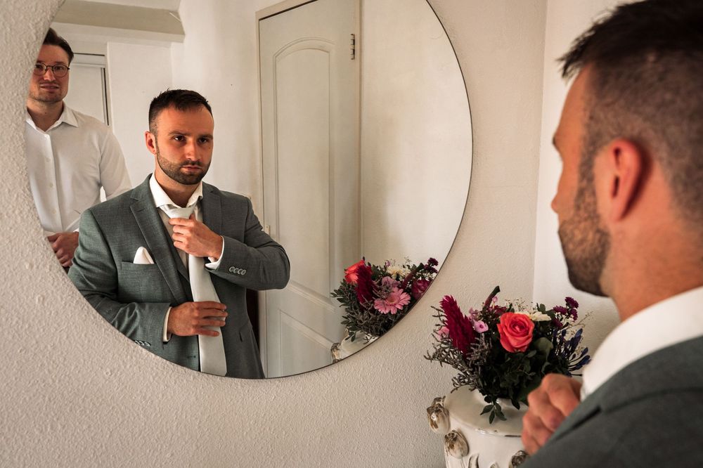 A person adjusts their tie while looking in a circular mirror during wedding preparations.