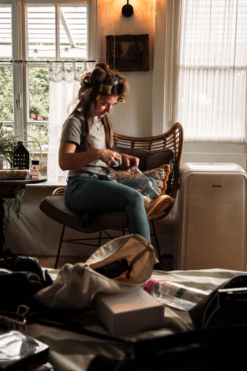 A cozy morning scene with someone in hair curlers sitting by a window in a modern chair with scattered items nearby.
