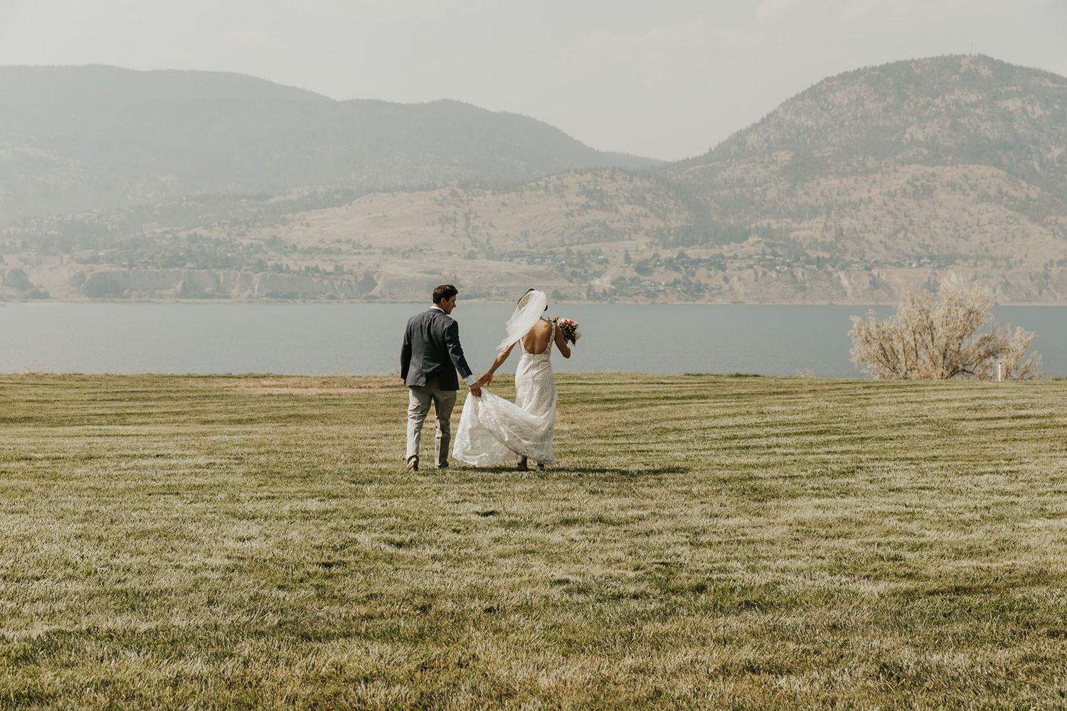 Bride and groom walking hand-in-hand across green meadow with scenic lake and mountain backdrop.