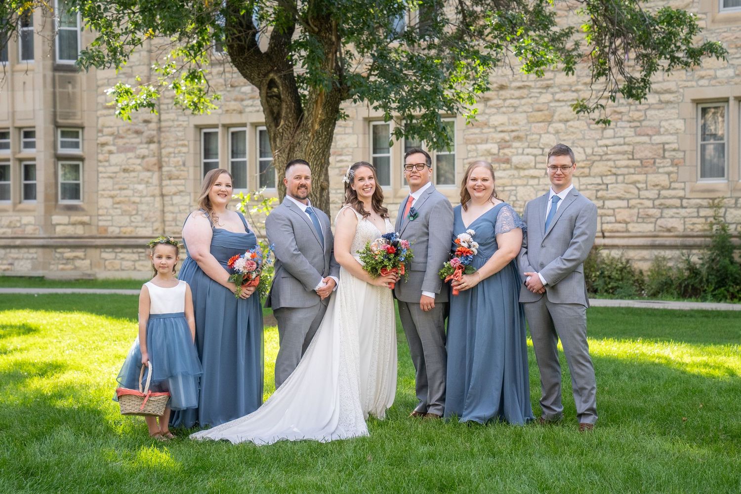 Wedding party in gray suits and a white dress pose together on a sunny lawn near a stone building.