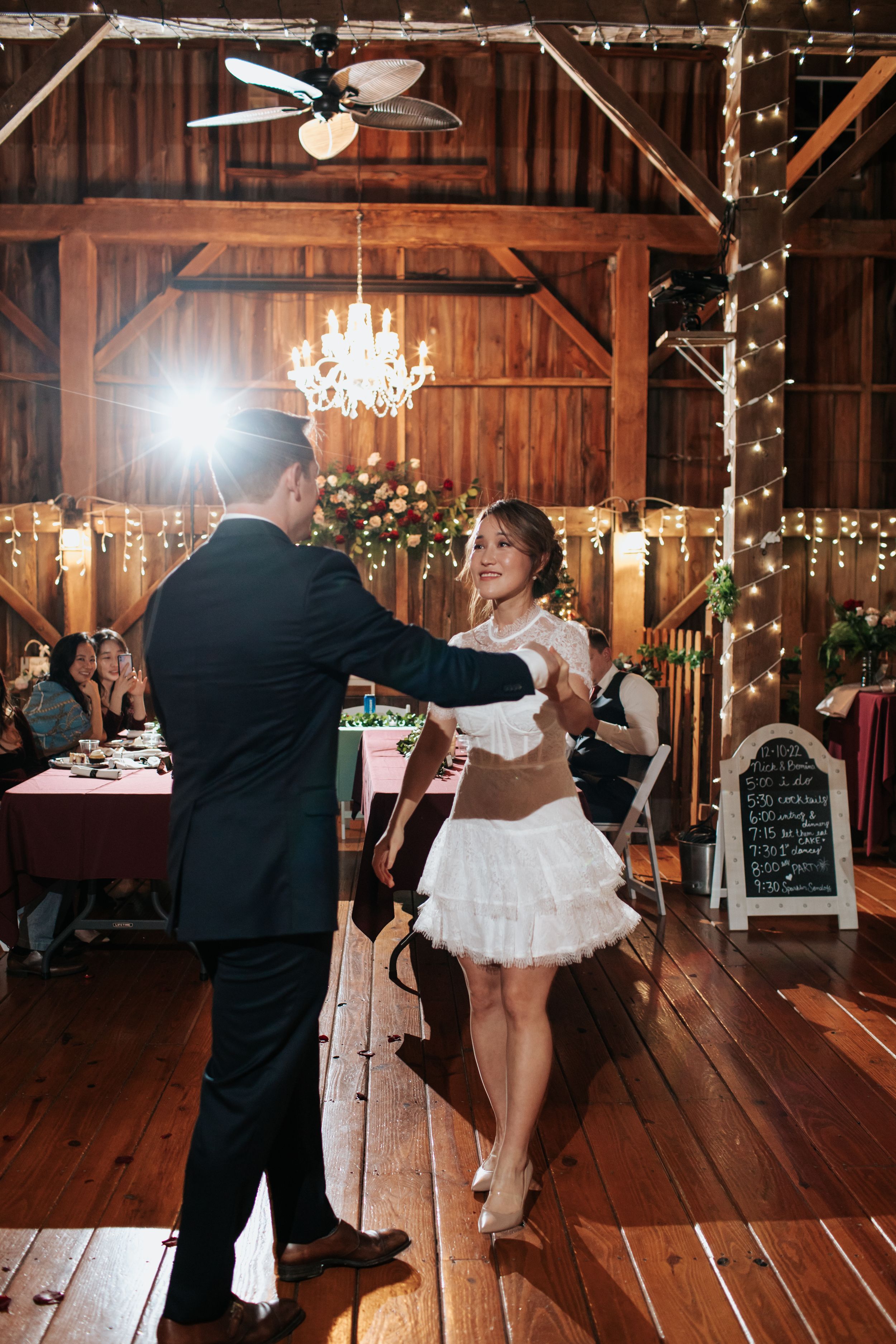 A couple shares a romantic first dance in a rustic barn wedding venue decorated with twinkling lights and chandeliers.