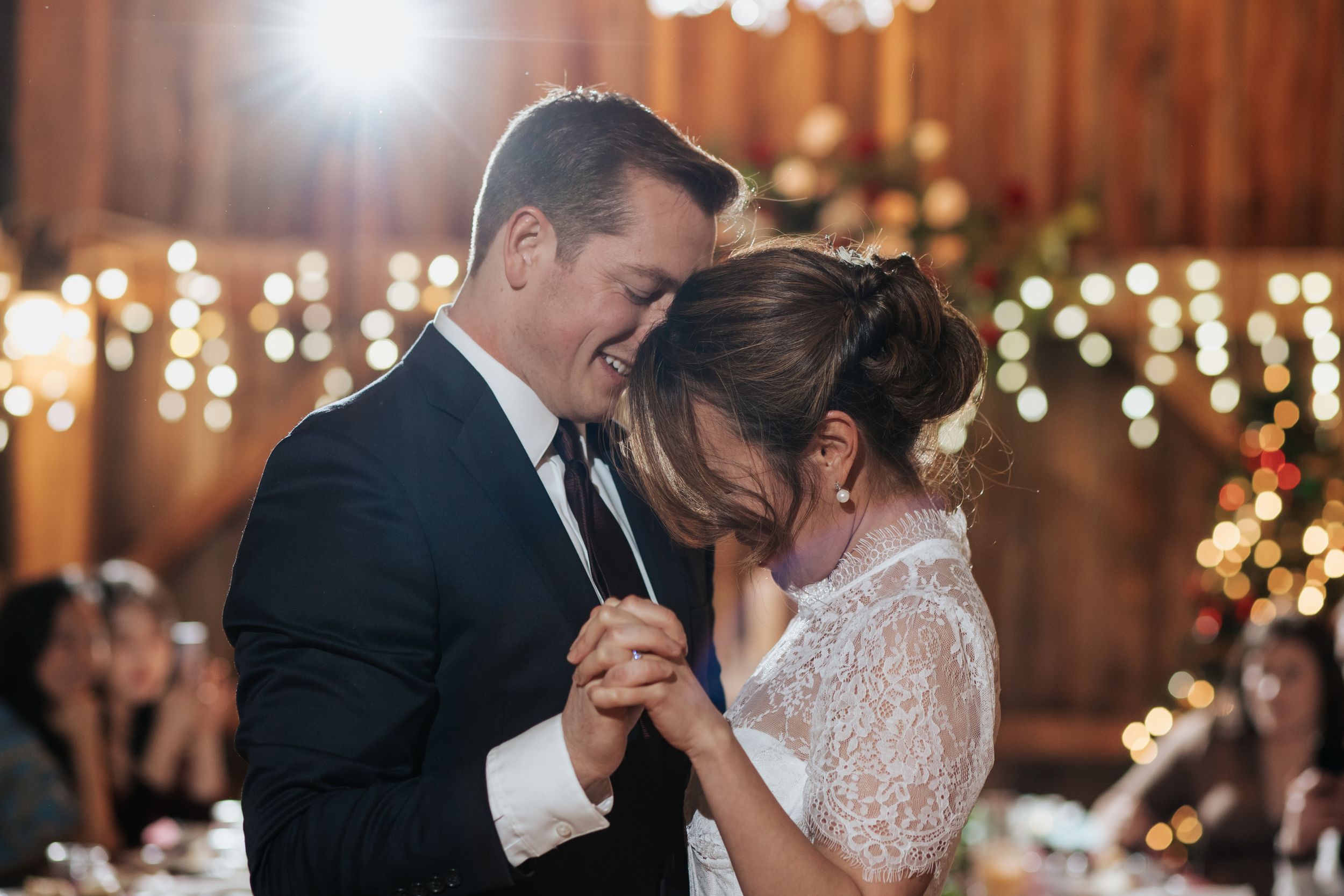 Newlyweds share an intimate dance under twinkling lights at a rustic barn wedding reception.