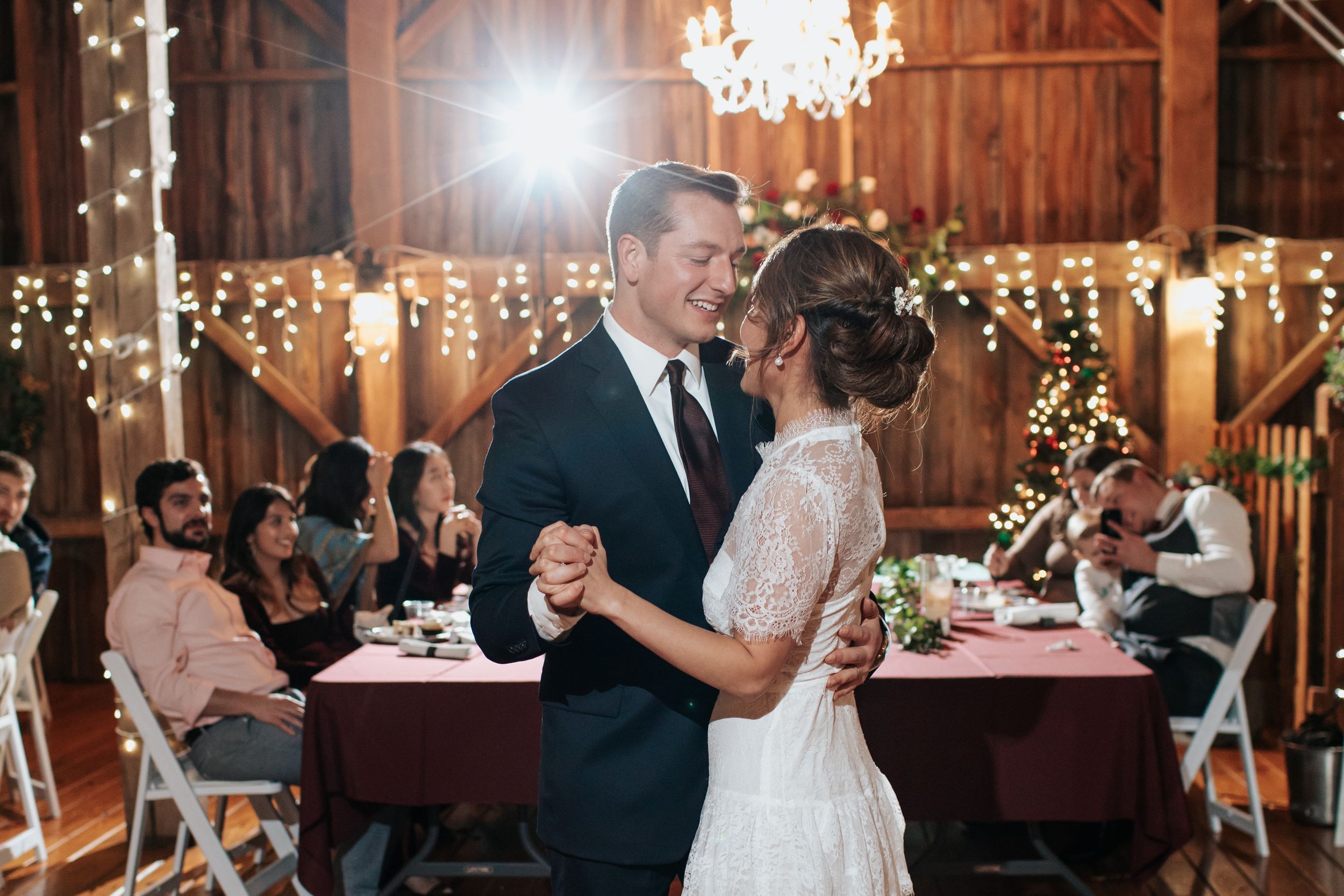Wedding guests dance and celebrate under twinkling lights in a rustic wooden barn venue during an evening reception.