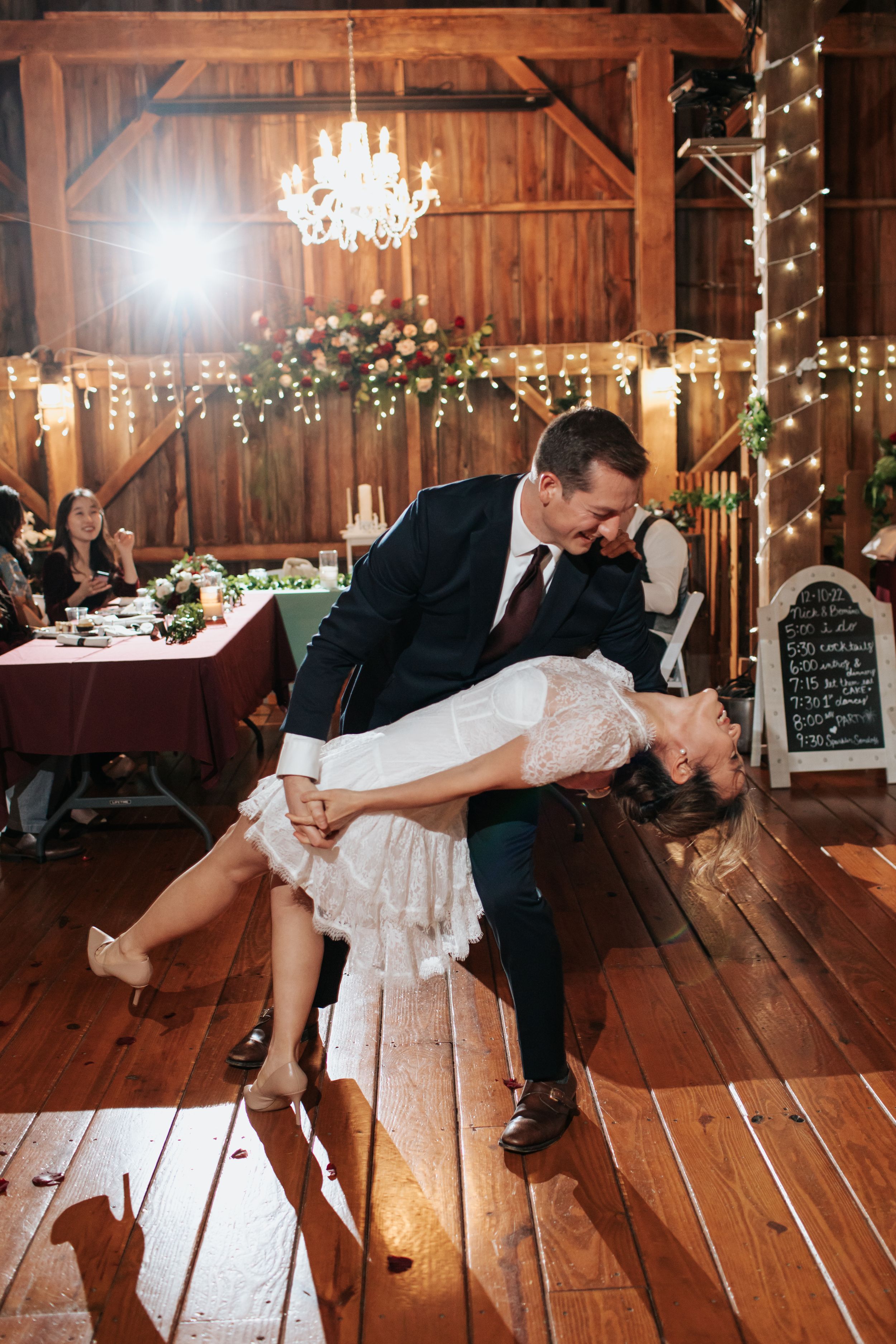 Romantic wedding couple dances and dips in rustic wooden barn decorated with twinkling lights and chandeliers.