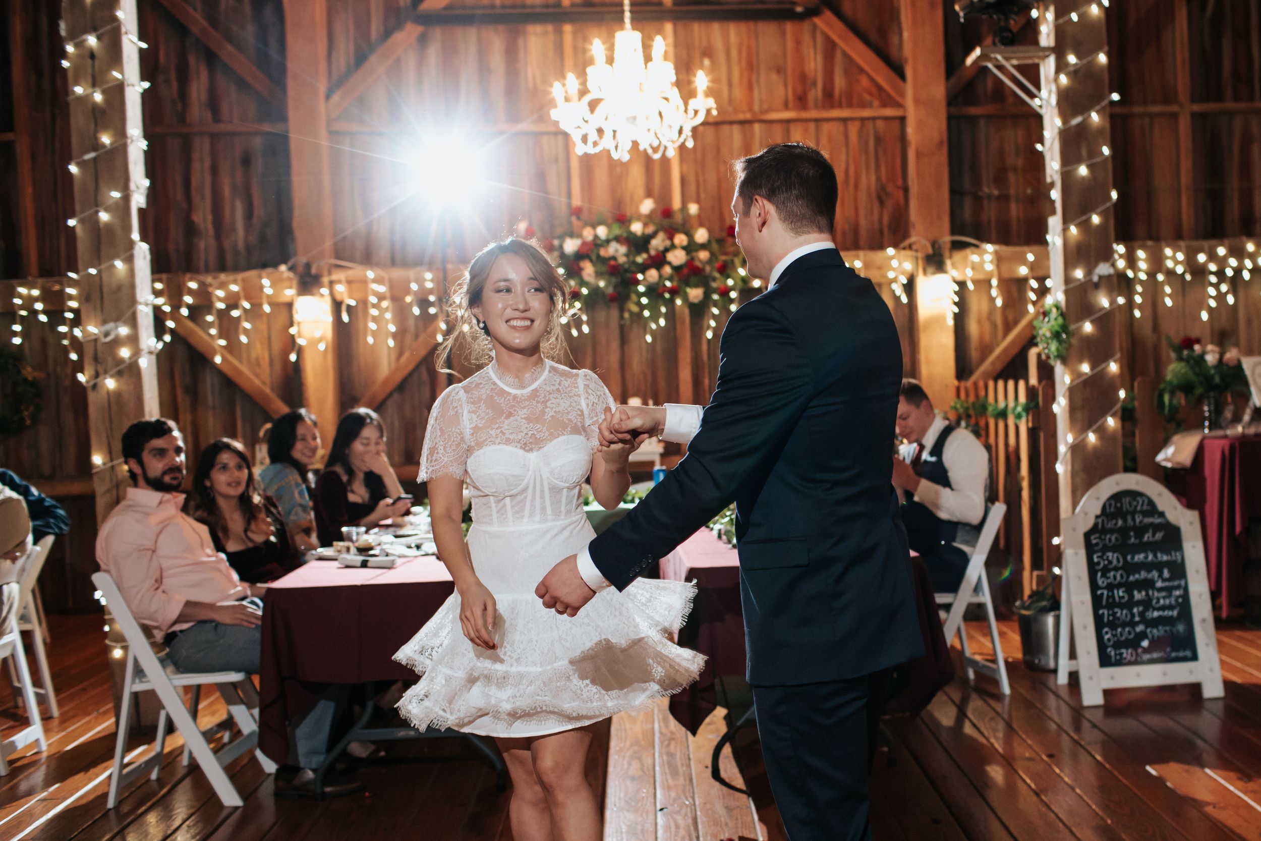 A couple shares their first dance in a rustic barn venue decorated with twinkling lights and an elegant chandelier.