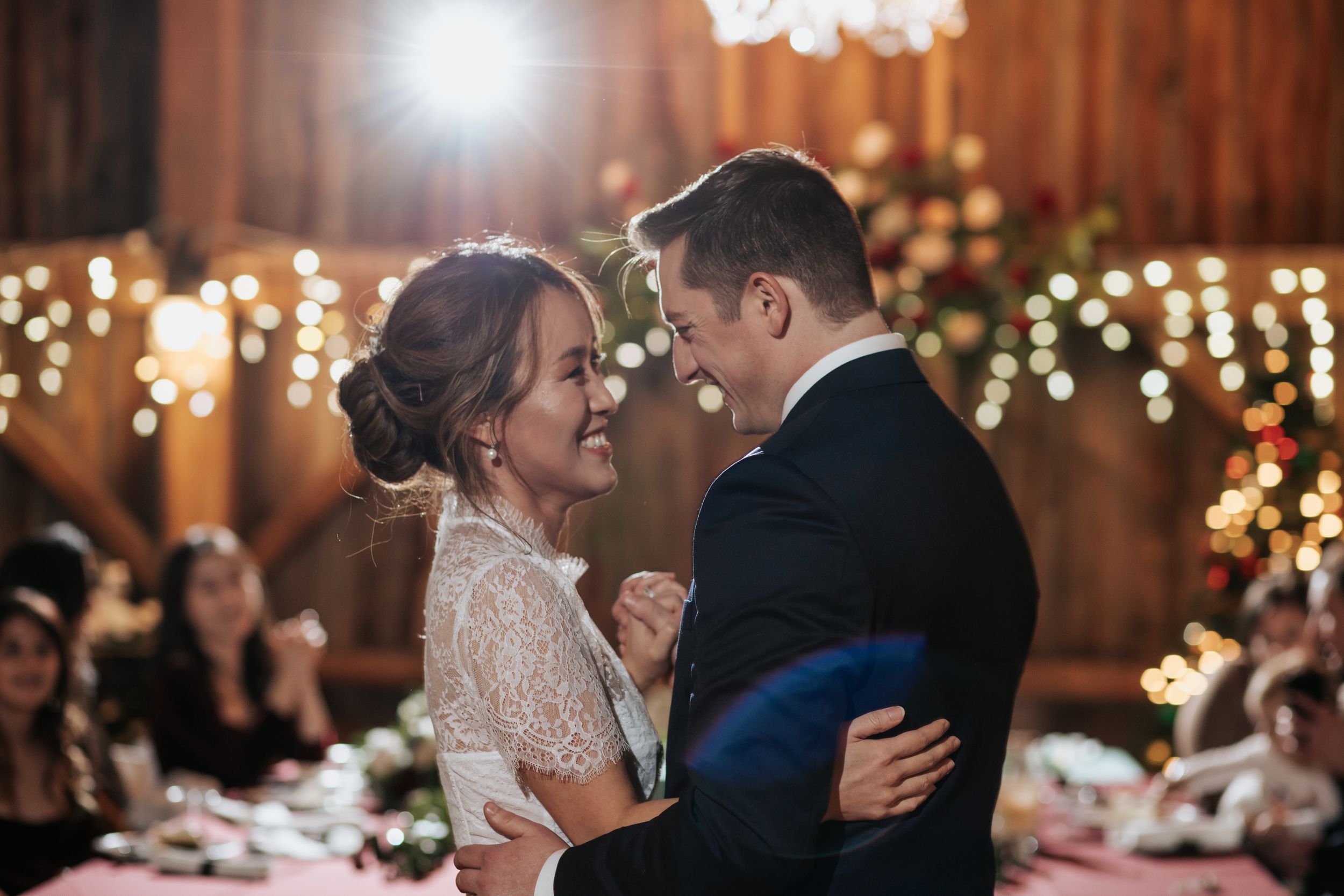 A romantic wedding reception dance moment in a rustic barn venue illuminated by warm string lights and chandeliers.