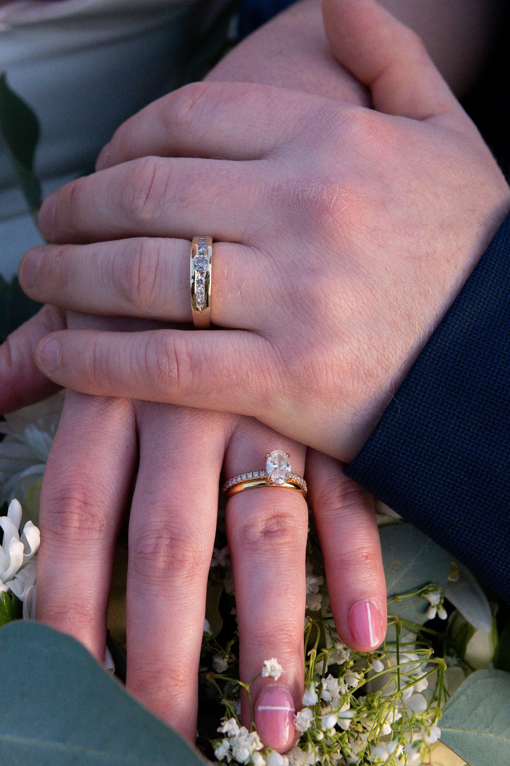 Two hands wearing wedding rings rest together on white flowers in a romantic pose.