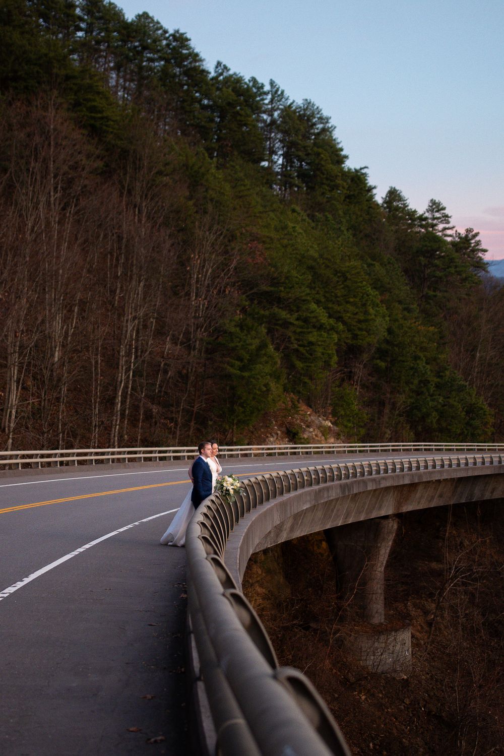 A curved mountain road with guardrails winds through a forested landscape at sunset.