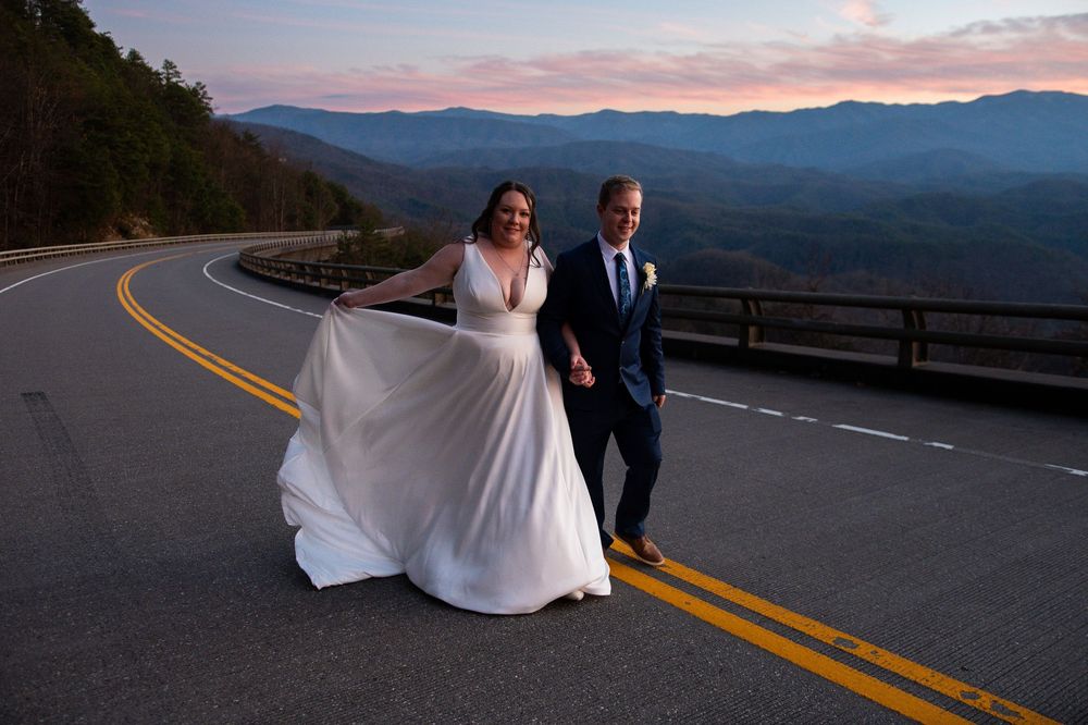 A couple poses on a scenic mountain road at sunset with the bride's white dress flowing in the wind.